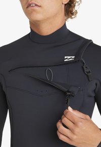 Traje de neopreno negro con cuello alto, que cuenta con un bolsillo con cremallera asimétrica y correas ajustables. Textura suave con costuras planas.