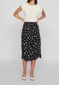 Kremowa bluzka o structurowanej fakturze z krótkimi falbanistymi rękawami, zestawiona z czarną spódnicą z rozproszonym kremowym wzorem w kwadraty, uzupełniona kremowymi balerinkami.