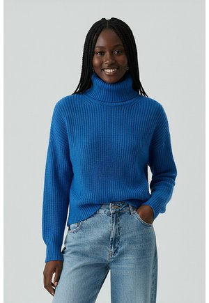 Blauer gerippter Rollkragenpullover mit langen Ärmeln, der eine lockere Passform und strukturierte Strickerei aufweist. Getragen mit hellblauen Jeans.