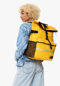 Mochila impermeable amarilla con correas negras, una franja reflectante y un bolsillo lateral de malla. El usuario lleva una camisa de patrón azul y unos vaqueros.