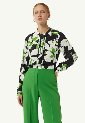 Bluse mit floralen Mustern in Schwarz, Weiß und Grün, mit langen Ärmeln und hohem Kragen, kombiniert mit grünen, hoch taillierten Hosen.