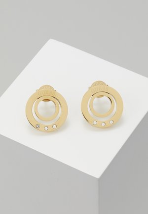 Boucles d'oreilles clous circulaires en or avec deux anneaux concentriques, chacun orné de trois petits cristaux blancs et "GUESS" gravé sur l'anneau supérieur.