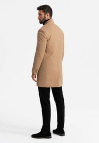 Cappotto su misura color beige con un design testurizzato, chiusura a un bottone e collo a pistagna, abbinato a pantaloni neri slim-fit e stivaletti alla caviglia.
