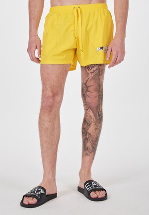 Bañador amarillo con cintura elástica y cordón. Presenta un logo impreso en el lado izquierdo. Acompañado de sandalias negras tipo playeras.