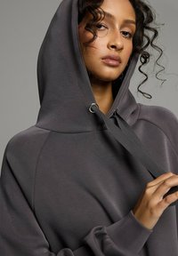 Ciemnoszara bluza z kapturem z kieszenią typu kangur, kapturem ze sznurkiem i ściągaczami przy rękawach. Miękka tekstura i luźny krój z widocznymi detalami szwów.