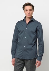 Camicia da uomo in tessuto verde scuro, con colletto classico, maniche lunghe e bottoni bianchi, abbinata a pantaloni grigi strutturati.