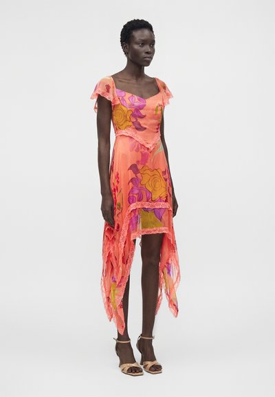 Γυναίκα που στέκεται φορώντας αμάνικο ασύμμετρο κοραλί φόρεμα με πορτοκαλί και μωβ ανθικά σχέδια και μπεζ σανδάλια με τακούνια.