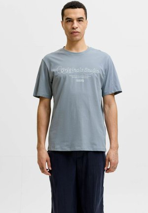 Mand med kort krøllet hår iført en grå T-shirt med teksten "Originals Studio" og mørke bukser, stående foran en ensfarvet hvid baggrund.