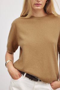 Trumpas rankovėmis megztinis su apvalia kaklo iškarpa, pagamintas iš minkštos karamelės spalvos medžiagos, derinamas su juodu diržu ir baltais chino kelnėmis. Lygus audinys, minimalistinis dizainas.