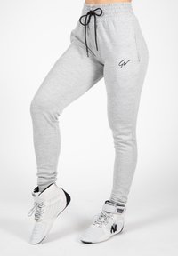 Light grey sweatpants with a drawstring waistband, featuring a black logo on the thigh and a tapered fit, paired with white athletic shoes.