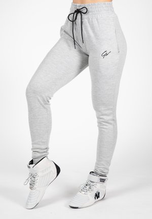 Light grey sweatpants with a drawstring waistband, featuring a black logo on the thigh and a tapered fit, paired with white athletic shoes.