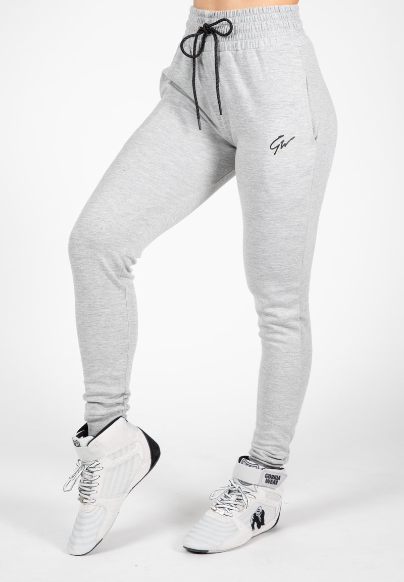 Light grey sweatpants with a drawstring waistband, featuring a black logo on the thigh and a tapered fit, paired with white athletic shoes.