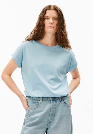 Femme aux longs cheveux bouclés portant un t-shirt bleu clair à manches retroussées, rentré dans un jean taille haute bleu clair, les mains dans les poches.