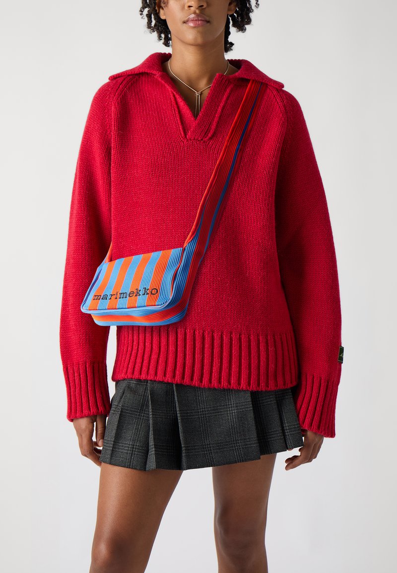 Red oversized knit sweater with a V-neck, ribbed cuffs, and hem. Paired with a black checkered pleated skirt and a striped blue, red, and orange bag.