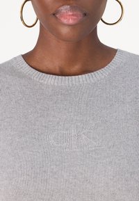Grå ribbad stickad tröja med rundad halsringning och en präglad "CK"-logotyp på bröstet. En slät textur och figurnära design förbättrar dess utseende.