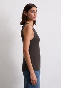 Frau mit schulterlangem dunklem Haar, die ein ärmelloses dunkelgraues Oberteil und blaue Jeans trägt und seitlich vor einer schlichten weißen Wand steht.