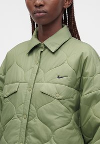 Prim-plan cu o persoană cu pielea închisă la culoare care poartă o geacă matlasată verde deschis cu nasturi capse și un mic logo negru Nike pe piept.