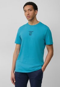Turquoise katoenen T-shirt met korte mouwen, ronde halslijn en zwarte geborduurde tekst "LONESOME RIDER SOCIAL Club" op de voorkant.