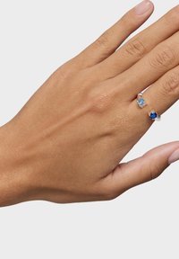 Srebrny pierścień z dwoma owalnymi kamieniami szlachetnymi: jasnoniebieskim kamieniem i ciemniejszym niebieskim kamieniem, na częściowo widocznej dłoni. Gładka faktura.