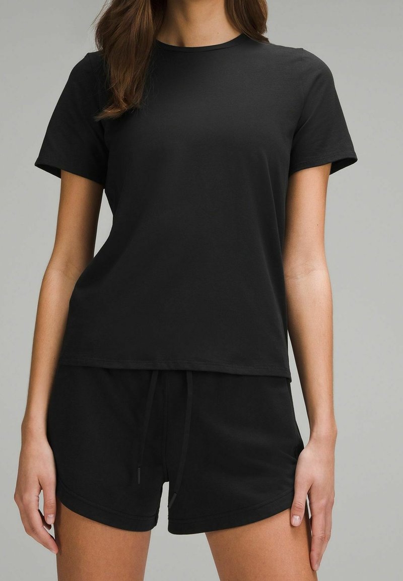 Femme portant un t-shirt noir uni à manches courtes et un short noir assorti, debout devant un fond neutre.