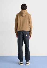 Mies, jolla on yllä ruskea huppari, mustat farkut, joissa brändipatchi, ja beiget tennarit, seisoo sinisellä lattialla valkoisen seinän edessä selin.