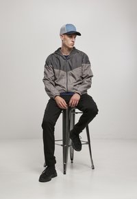 Grå og sort vindjakke med et geometrisk design, der sidder på en sølvfarvet barstol. Sorte bukser og sorte sneakers afslutter looket.