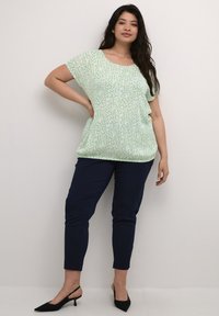 Maglietta verde chiaro con un motivo bianco testurizzato, abbinata a pantaloni slim-fit blu navy e décolleté nere con cinturino. Maniche corte, scollatura rotonda.
