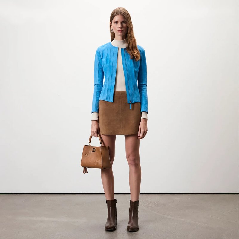 Blaue Lederjacke, cremfarbener Rollkragenpullover, brauner Wildleder-Minirock, braune Handtasche und braune Stiefeletten in einem minimalistischen Ambiente.