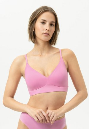 Jeune femme aux cheveux courts bruns portant un soutien-gorge rose clair et une culotte assortie sur un fond blanc uni.