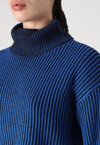 Blå og svart stripete genser med høy hals, laget med ribbestruktur og avslappet passform.