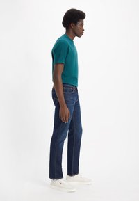 Tyrkise bomull t-skjorte kombinert med mørkeblå denimjeans. Modellen har på seg hvite sneakers og står i profil mot en ensfarget bakgrunn.