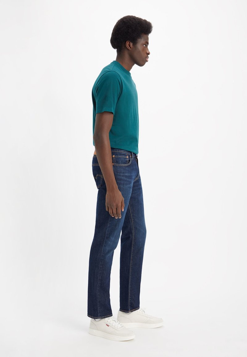 Camiseta de algodón color verde azulado combinada con jeans de mezclilla azul oscuro. El modelo lleva zapatillas blancas y está de perfil contra un fondo plain.