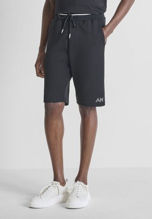 Hombre con pantalones cortos negros con cordón y logo en la pierna izquierda, y zapatillas blancas, de pie con una mano en el bolsillo sobre un fondo liso.