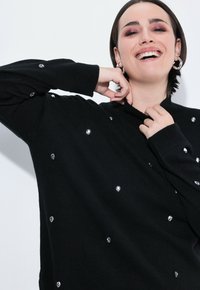 Maglione nero lavorato a maglia con collo alto e dettagli in gemme trasparenti sparse, che creano un aspetto testurizzato con maniche a costine.