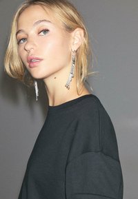 Orecchini a chandelier argentati con catene a strati e accenti circolari, indossati con una felpa nera con scollo rotondo e maniche corte.