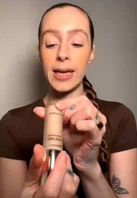Femme aux cheveux tressés tient une bouteille de MAC Skinfinish Compact Foundation et en indique les caractéristiques en les expliquant.