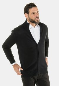Sort strikket zip-up cardigan med ribbet krave og manchetter, der har sidelommer og en glat tekstur, båret over en hvid skjorte.