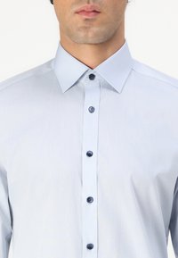 Világoskék, sima anyagból készült ing. Elegáns, hegyes gallérral és sötétkék gombokkal rendelkezik az elején.