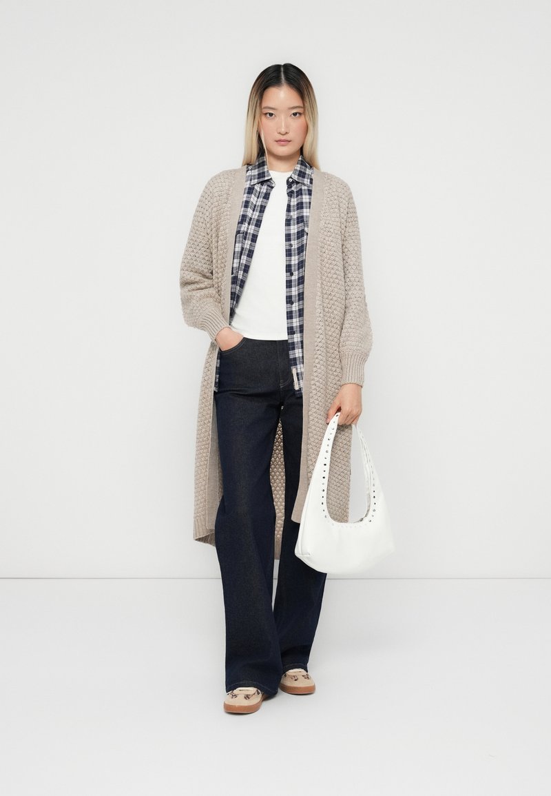 Cardigan en tricot beige, chemise à carreaux bleu marine, t-shirt blanc, jeans foncés et chaussures à enfiler beiges. Sac blanc avec un détail texturé.