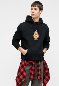 Μαύρη φόρμα με κουκούλα, με κεντρικό γραφικό φλόγας και κείμενο "SANTA CRUZ". Ύφασμα από βαμβάκι, με τσέπη καγκουρό, φορεμένη με κόκκινο και μαύρο καρό πουκάμισο δεμένο στη μέση.