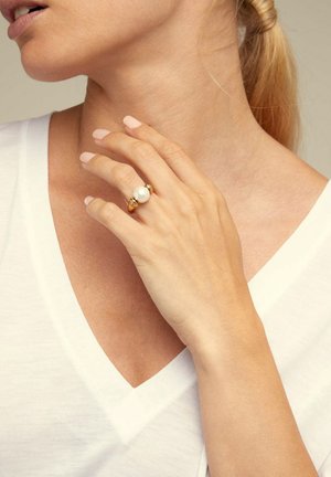Anillo de oro con una gran perla blanca y dos acentos dorados más pequeños, lucido en una mano con uñas naturales y claras. Top blanco de escote en V.