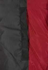 Giacca imbottita con materiale esterno nero e fodera rossa, con una tasca visibile sul lato sinistro. Tessuto testurizzato e resistente all'acqua.
