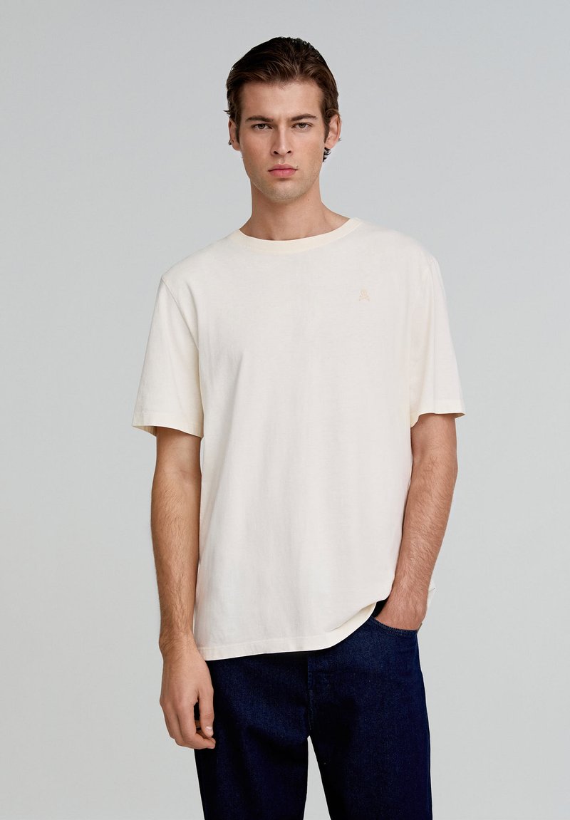 Camiseta de algodón color crema con cuello redondo, mangas cortas y un pequeño detalle del logo en el pecho. Modelo lleva puestos unos jeans azules.