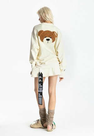 Mujer con cabello rubio que lleva una sudadera oversize de color crema con una gran cara de oso de peluche en la espalda, una falda a juego y zapatos beige con calcetines.