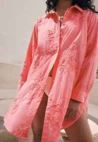 Rožinė siuvinėta marškinėliai su ilgomis rankovėmis, puošta gėlių raštais ir sagomis priekyje; laisvo kirpimo ir lengvos medžiagos.