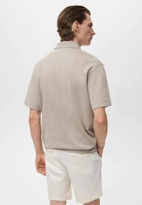 Beige kortärmad pikétröja med en standardkrage, slät textur. Kombinerad med vita shorts, sett bakifrån. Minimalistisk design.