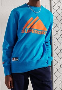Sudadera color turquesa con un gran logo naranja de "SUPERDRY", puños y dobladillo de canalé, fabricada en un suave material de felpa. Presenta una etiqueta de marca en la parte inferior.
