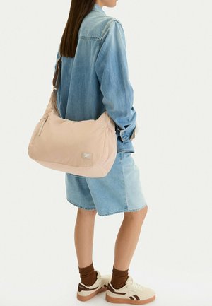 Persona con giacca di jeans e pantaloncini che porta una borsa a tracolla beige Reebok, indossando sneakers bianche con calzini marroni, in piedi su sfondo bianco.