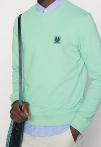 Mintgrön sweatshirt med ribbmuddar och nertill, med en rektangulär logga patchad på bröstet. Bärs över en ljusblå skjorta med krage.