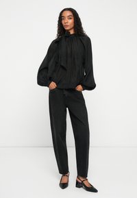 Black satin blouse with a high neck and tie detail, paired with high-waisted black denim jeans and black heeled pumps.